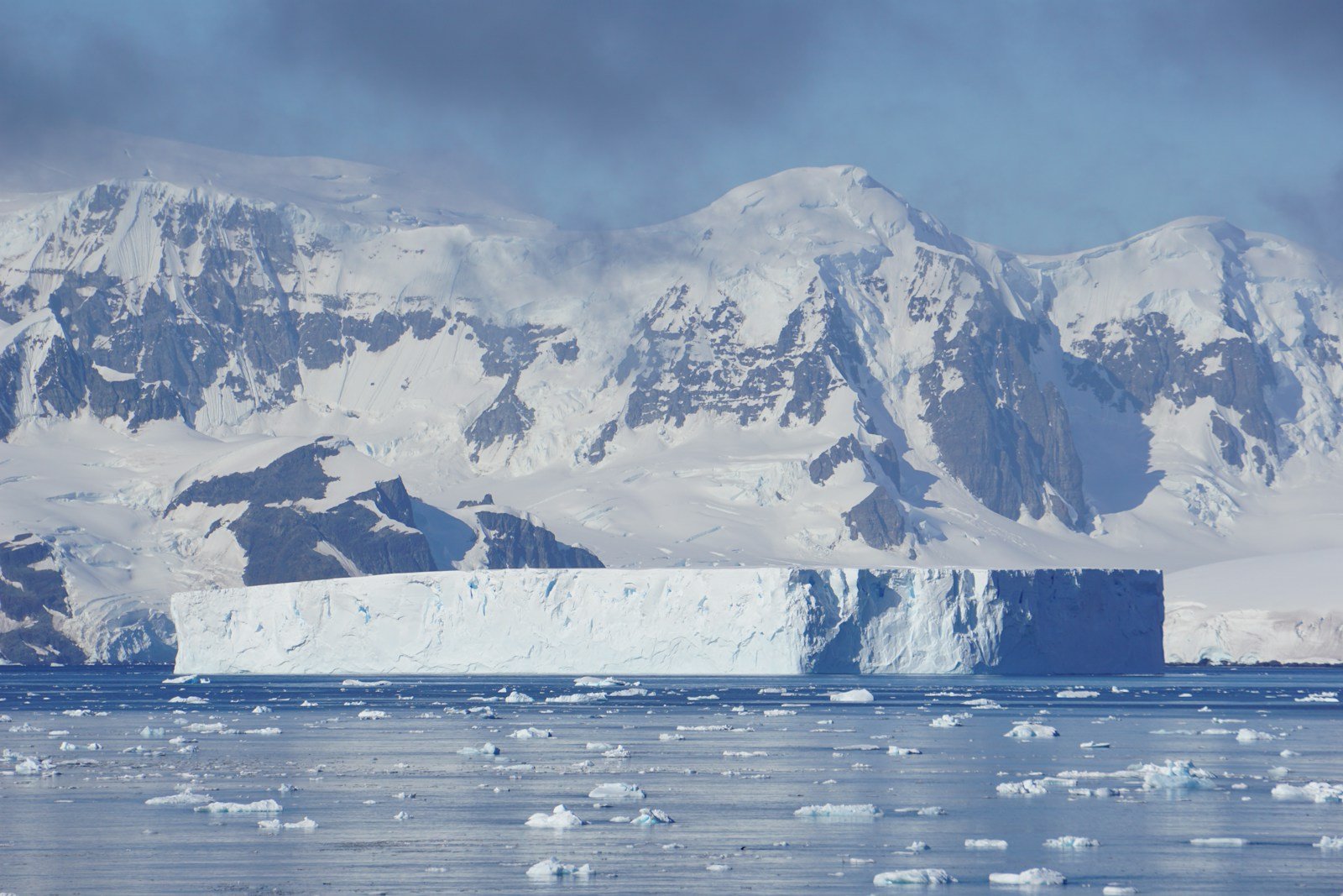 An iceberg floats in front of snowy mountains.